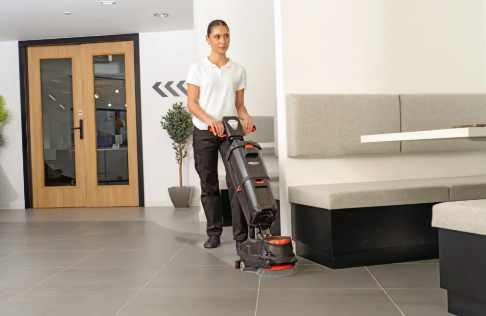 A woman operating the Numatic 220NX floor scrubber on a grey tiled floor in a modern building lobby, leaving a clean path.