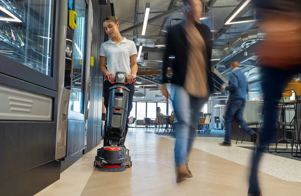 A woman cleaning a corridor in a busy commercial lounge with a Numatic 220NX floor scrubber, while people walk past.