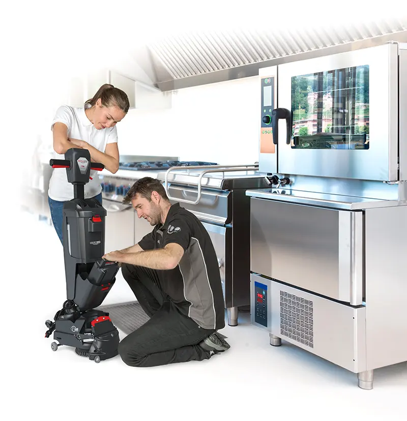 A Numatic floorcare expert kneels to demonstrate a feature of the 220NX floor scrubber dryer to a woman in a professional kitchen, as part of the on-site training.