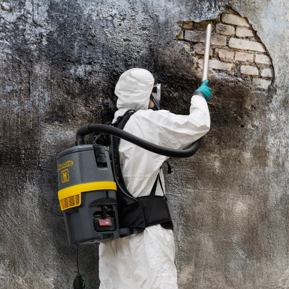 Worker in full hazmat and respirator using a Numatic RHB150NX H-Class vacuum to clean dust from a dilapidated wall.